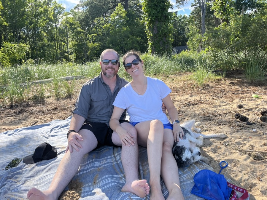 Liz and Mike sitting on a blanket at the beach with their dog.